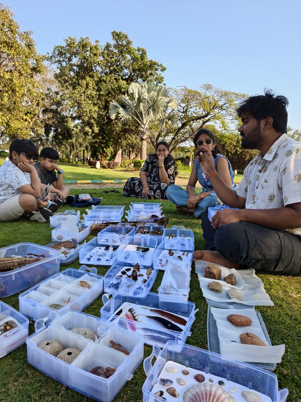 Families sit on the grass in a park around open specimen trays as the educator holds up a seed
