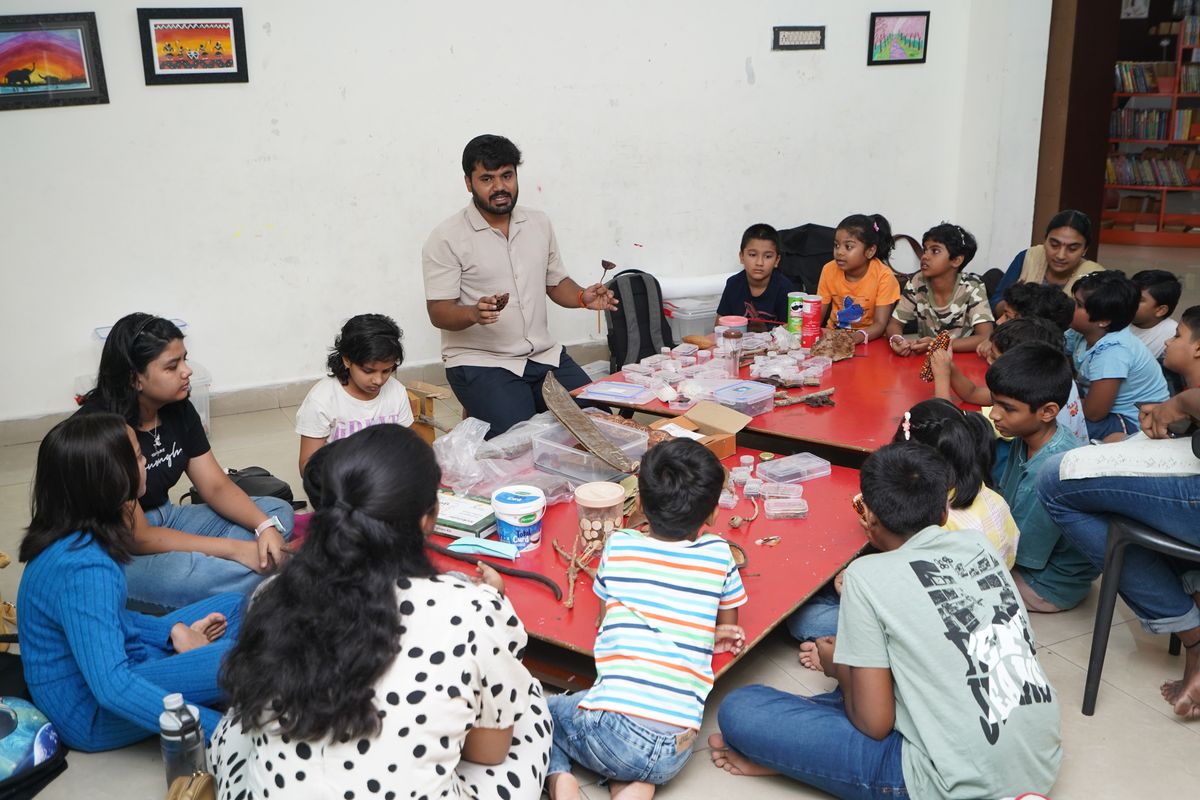Children sit around low red tables in a library as the educator shows them seed pods